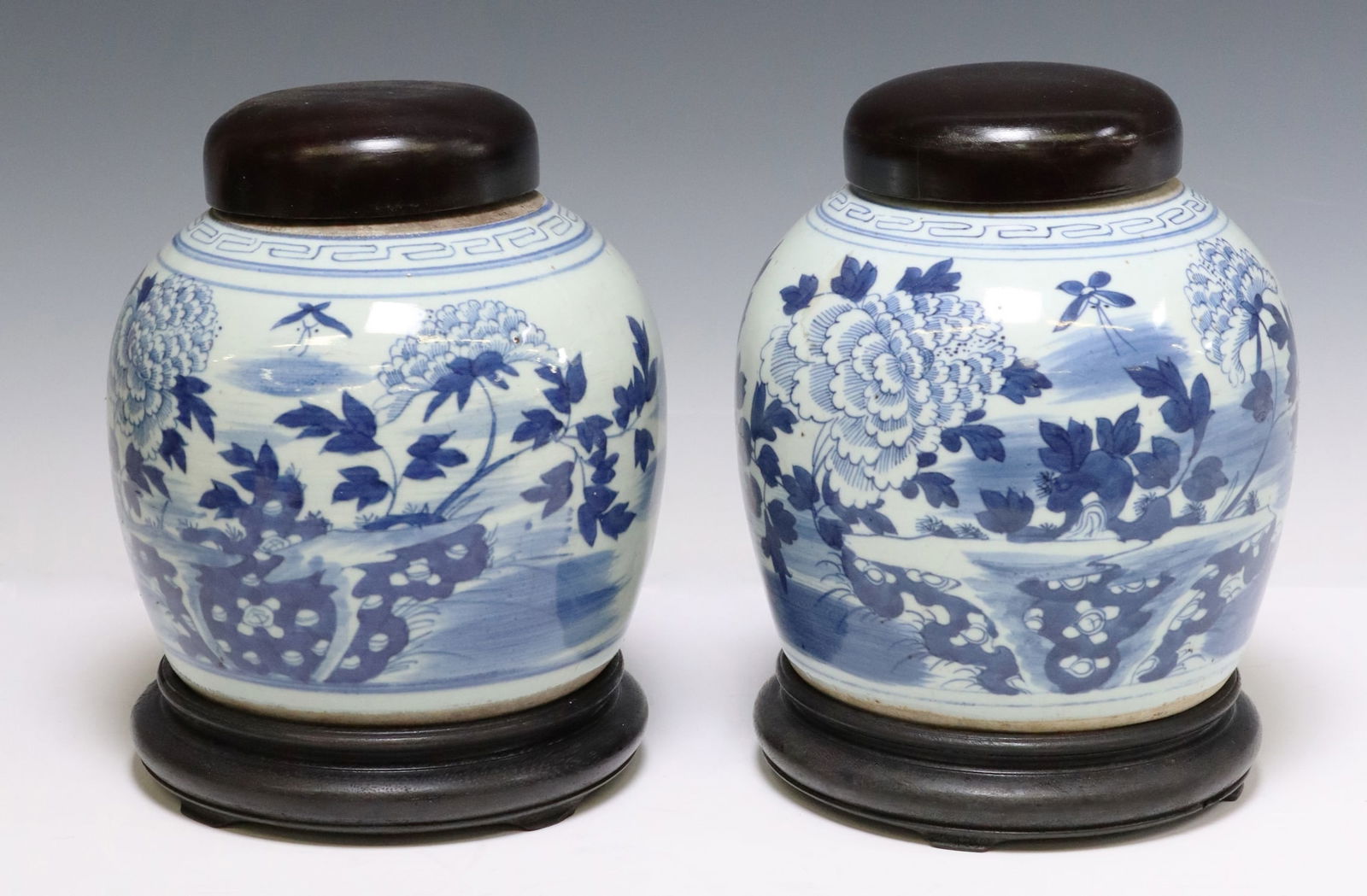 (2) CHINESE BLUE & WHITE PORCELAIN GINGER JARS: (pair) Chinese blue and white porcelain ginger jars, each having wood lid, over tapered body with floral landscapes, meander trim, rising on wood stand, jar: approx 8.5"h, 7"diam, on base: approx 10.5
