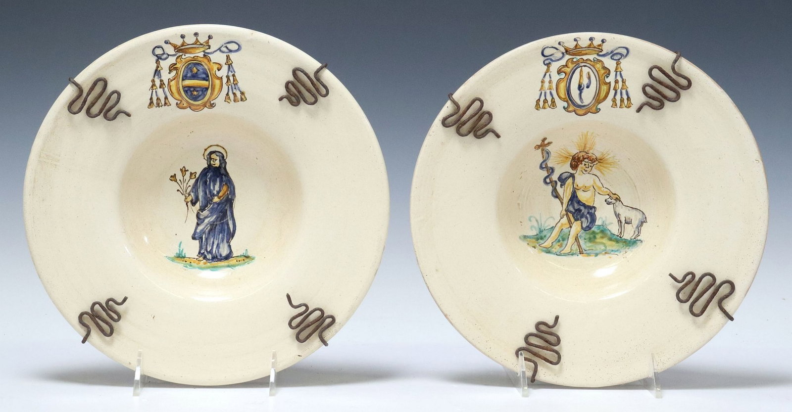 (2) POTTERY FOOTED BOWLS MADONNA & ST JOHN THE BAPTIST: (lot of 2) Tin-glazed earthenware footed bowls, each with an armorial bearing, including: (1) depicting the Madonna, (1) the Infant Saint John the Baptist with a lamb, each mounted in a wrou