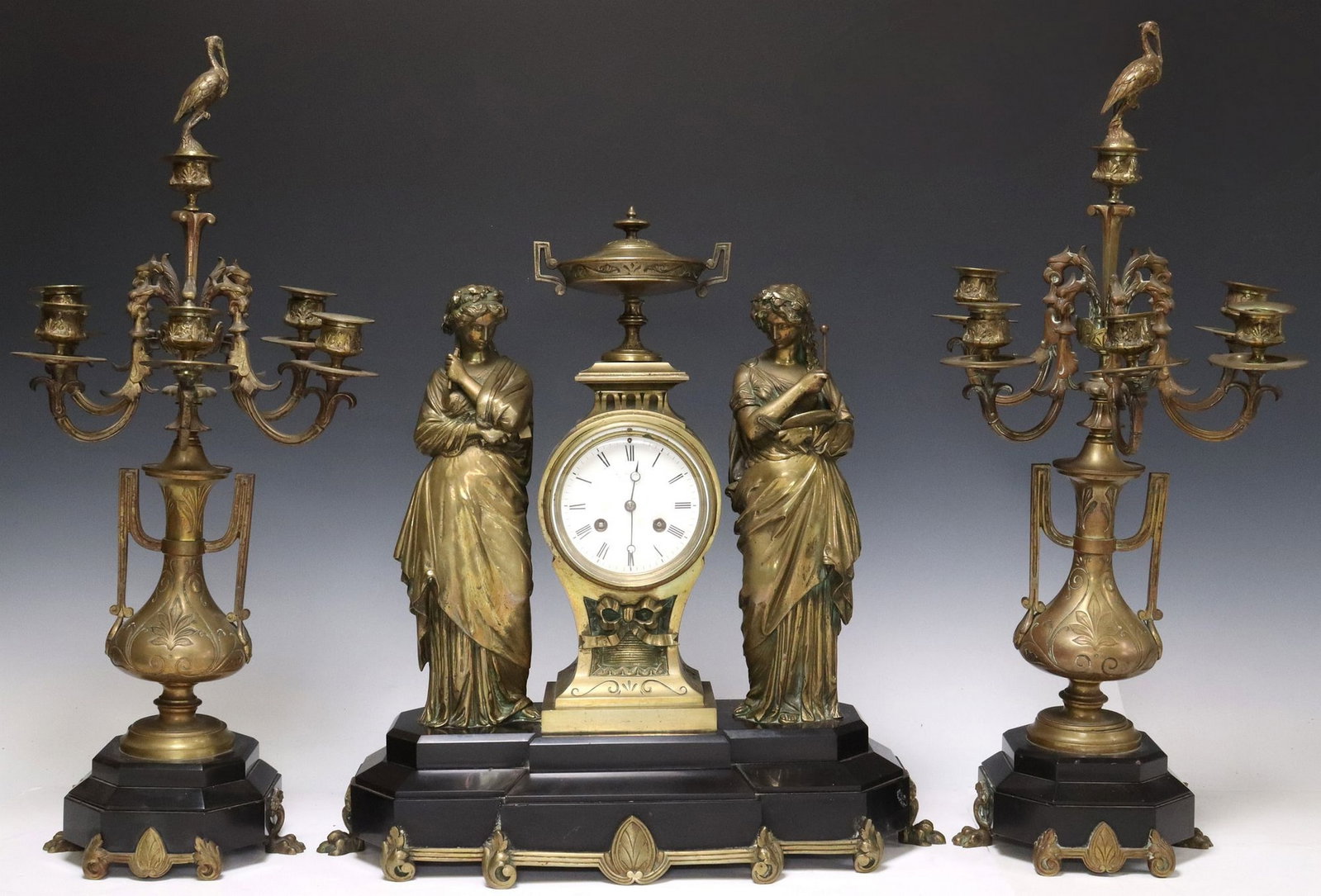 (3) FRENCH FIGURAL BRONZE MANTEL CLOCK & CANDELABRA GARNITURES: (lot of 3) French bronze mantel set, late 19th c., each piece on stepped slate base, further raised on paw feet, including: (1) mantel clock, having tazza-form topper flanked by two Classical sty