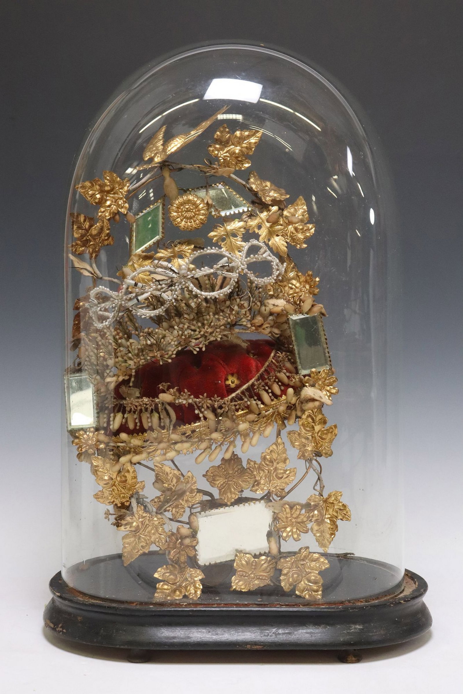 FRENCH GILT METAL GLOBE DE MARIEE/ BRIDE'S GLOBE: French globe de mariee/ bride's globe, late 19th c., glass cloche/ dome on ebonized base, enclosing gilt metal frame, beveled mirrors and floral garland encircling tufted red velvet cushion, cente