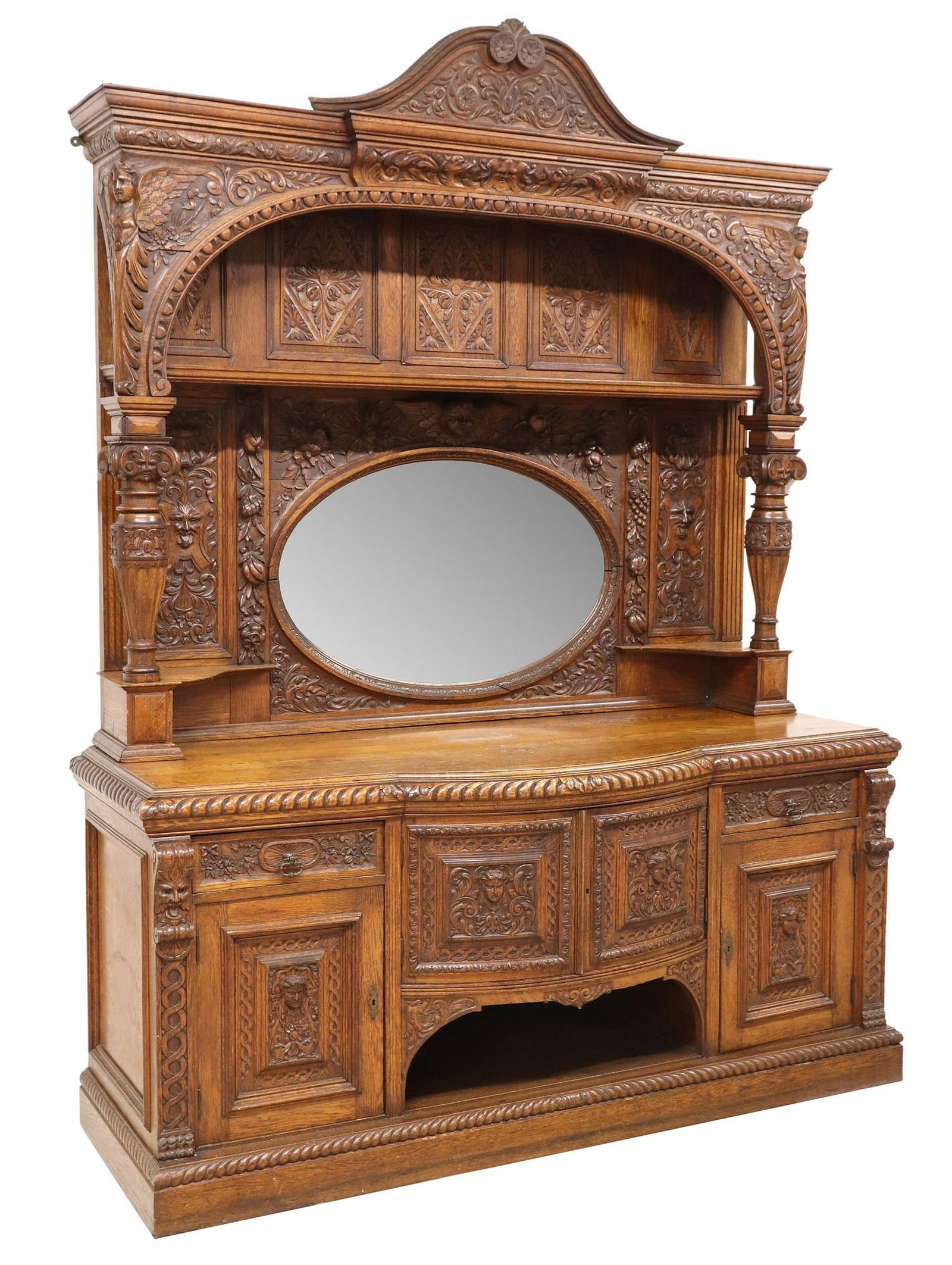 MONUMENTAL ENGLISH CARVED OAK MIRRORED SIDEBOARD: Monumental English carved oak sideboard,, early 20th c., swan's neck pediment, open display shelf behind an arcaded surround with winged angel corner posts, carved backboard with masks and fruit penda