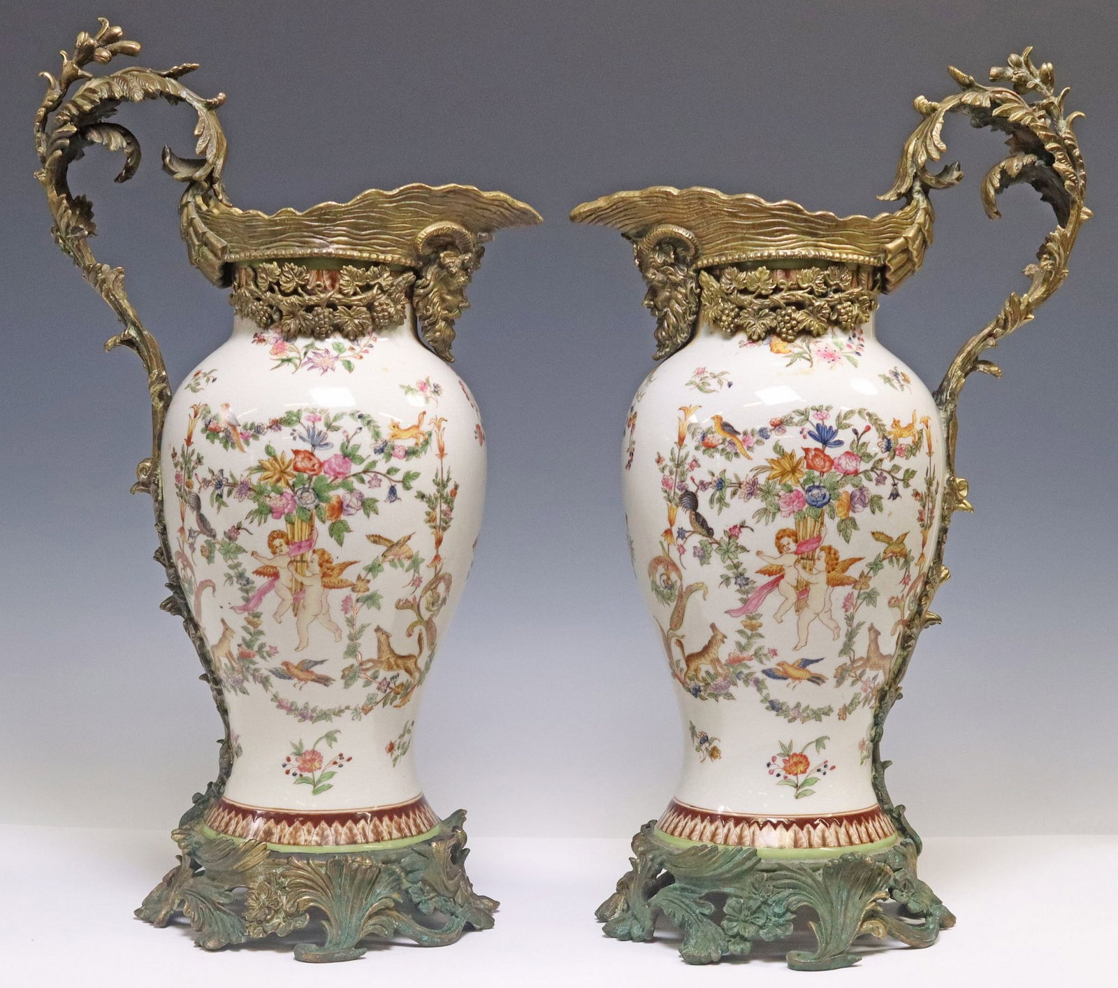 (2) NEOCLASSICAL STYLE BRONZE-MOUNTED PORCELAIN EWERS: (pair) Neoclassical style bronze-mounted porcelain ewers, late 20th c., each having scrolled foliate handle with figural mask and grape vine motifs, tapered porcelain body depicting cherubs and floral