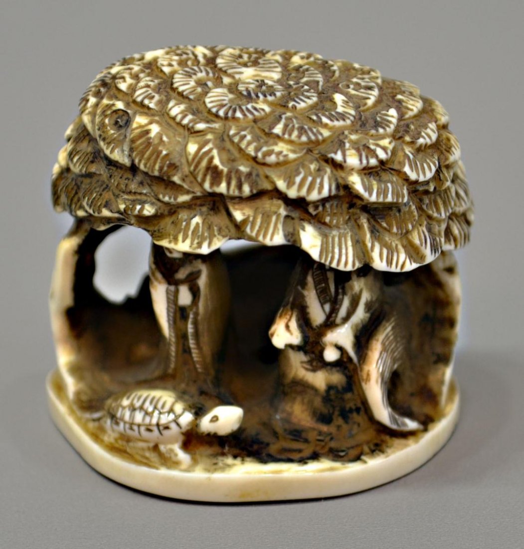 JAPANESE CARVED FIGURAL IVORY NETSUKE: *International shipping is not available on this lot* Japanese ivory netsuke, carved as an imaginary landscape with a turtle and a pair of figures sheltered by a large pine, the base signed [Moto]tomo