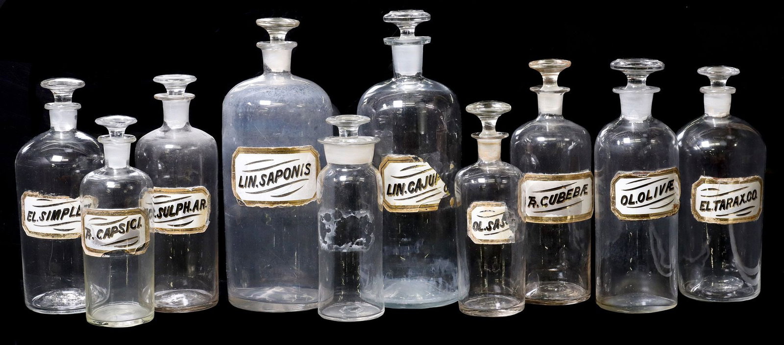 (10) ANTIQUE COLORLESS GLASS APOTHECARY / PHARMACY BOTTLES: (lot of 10) Antique colorless glass pharmacy or apothecary bottles, 19th c., each with parcel gilt ingredient labels, highlights include: (1) Lin. Saponin, (1) TR. Capsici, (1) El. Tarax.CO.