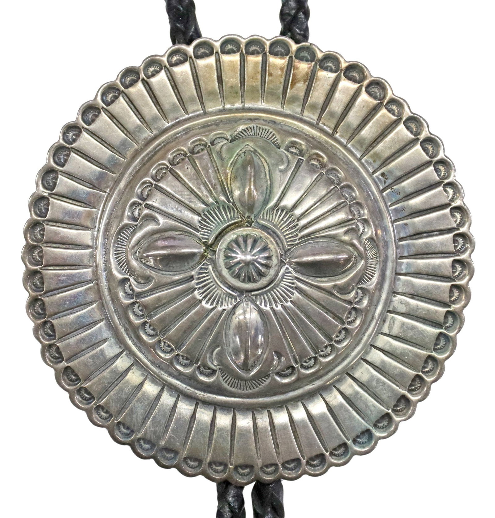 WILBERT SECATERO NAVAJO STERLING SILVER CONCHA BOLO TIE: Native American sterling silver bolo tie, signed WS. (Wilbert Secatero, Navajo/ Dine, 20th c.), with radiating stampwork motifs forming central floral medallion, silver tips, on braided black leather 