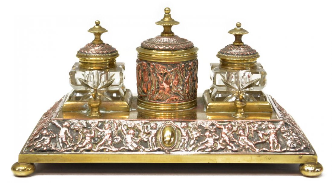 ANTIQUE CONTINENTAL BRASS & COPPER PUTTI INKSTAND: Continental brass and copper inkstand, 19th c., ornate scenes of putti amid grapevines in relief on the stand, fitted with a pair of square glass ink bottles, central covered receptacle of circular fo