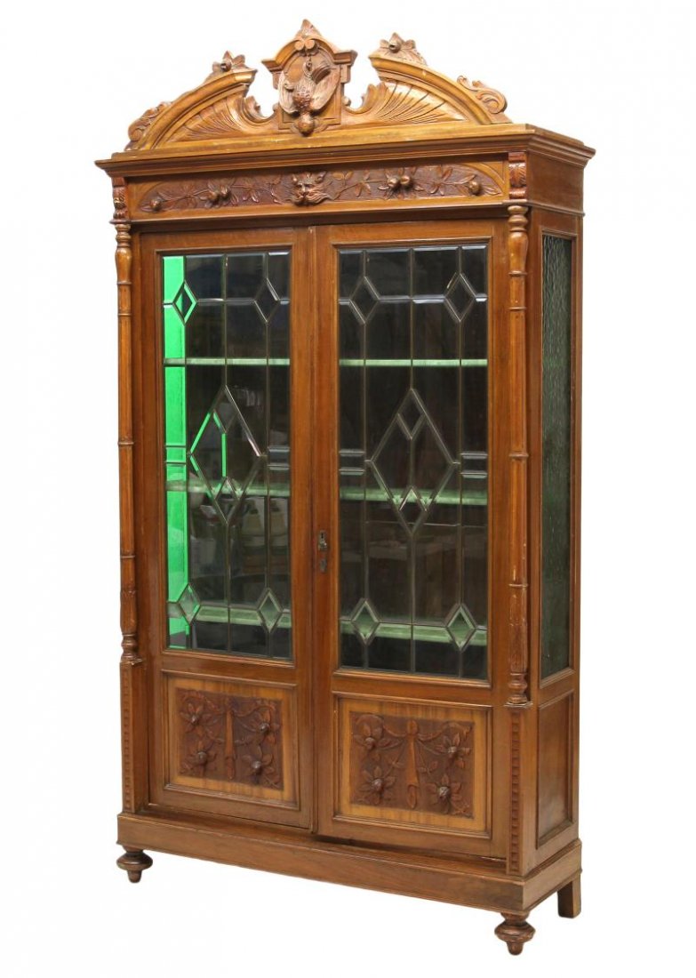 ITALIAN RENAISSANCE REVIVAL LEADED GLASS BOOKCASE: Italian Renaissance Revival walnut bookcase, late 19th c., an arched split pediment cornice over foliate and mask carved frieze, columns flanking two long doors with geometric shaped leaded beveled gl