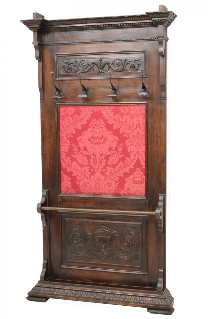 ITALIAN RENAISSANCE REVIVAL CARVED WALNUT HALLTREE: Italian Renaissance Revival walnut hall tree, late 19th c., carved cornice above four floriform coat / hat hooks, centered crimson foliate fabric and foliate carved panel enclosing a metal umbrella ra