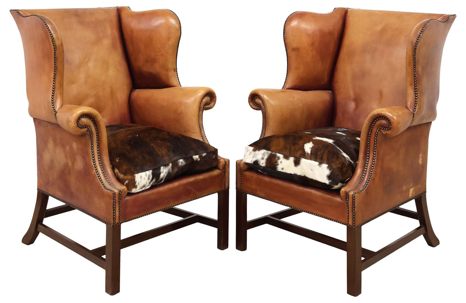 (2) ENGLISH LEATHER & COWHIDE UPHOLSTERED WINGBACK ARMCHAIRS: (pair) English Edwardian period armchairs, early 20th c., in a worn tan leather upholstery, bolster wings and arms, finished with nail head trim, removable seat cushion in a cowhide upholstery, rising