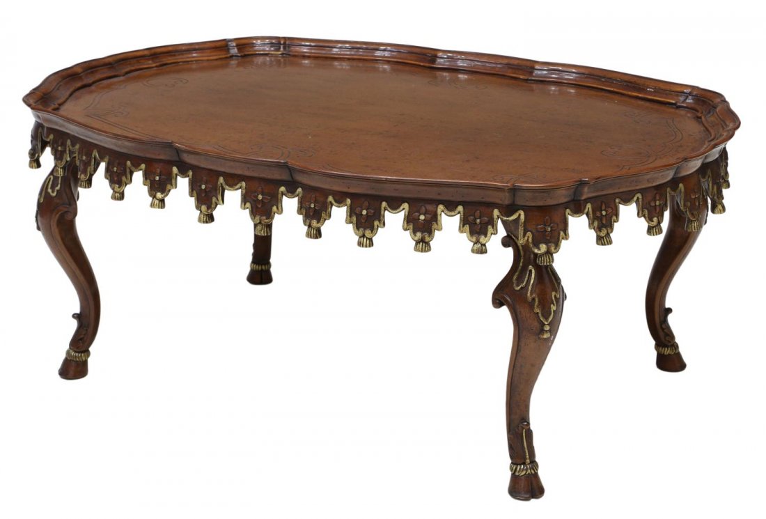 LARGE CARVED OVAL COFFEE TABLE: Large oval coffee table, 20th c., mahogany finish, the top with a stepped scalloped edge, incised carving, the skirt with gilt accented carved wood tassels, rising on cabriole legs with hoof feet, 20"