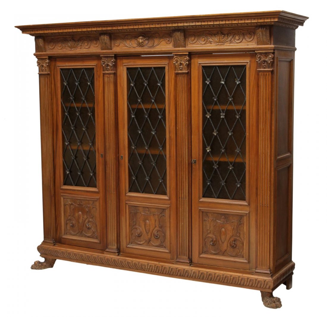 ITALIAN RENAISSANCE REVIVAL TRIPLE BOOKCASE: Italian Renaissance Revival walnut bookcase, early 20th c., having a stepped protruding cornice, a mask and foliate carved frieze, three partial glass front doors with wrought iron grills, all rising 