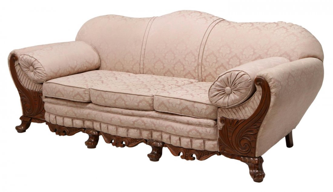AMERICAN VICTORIAN CAMEL BACK SOFA: American Victorian camel back sofa, c. 1900, upholstered in light pink fabric with white foliate accents, three cushions with bolsters on either end, the skirt and arm fronts with carved scrolled waln