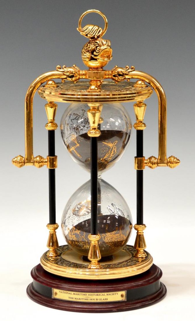 MARITIME FRANKLIN MINT HOURGLASS SAND TIMER: National Maritime Historical Society hourglass sand timer, Franklin Mint, glass case with gilt zodiac animals decor enclosed by an ornate brass frame with fish finial, accompanied by labeled wooden ba
