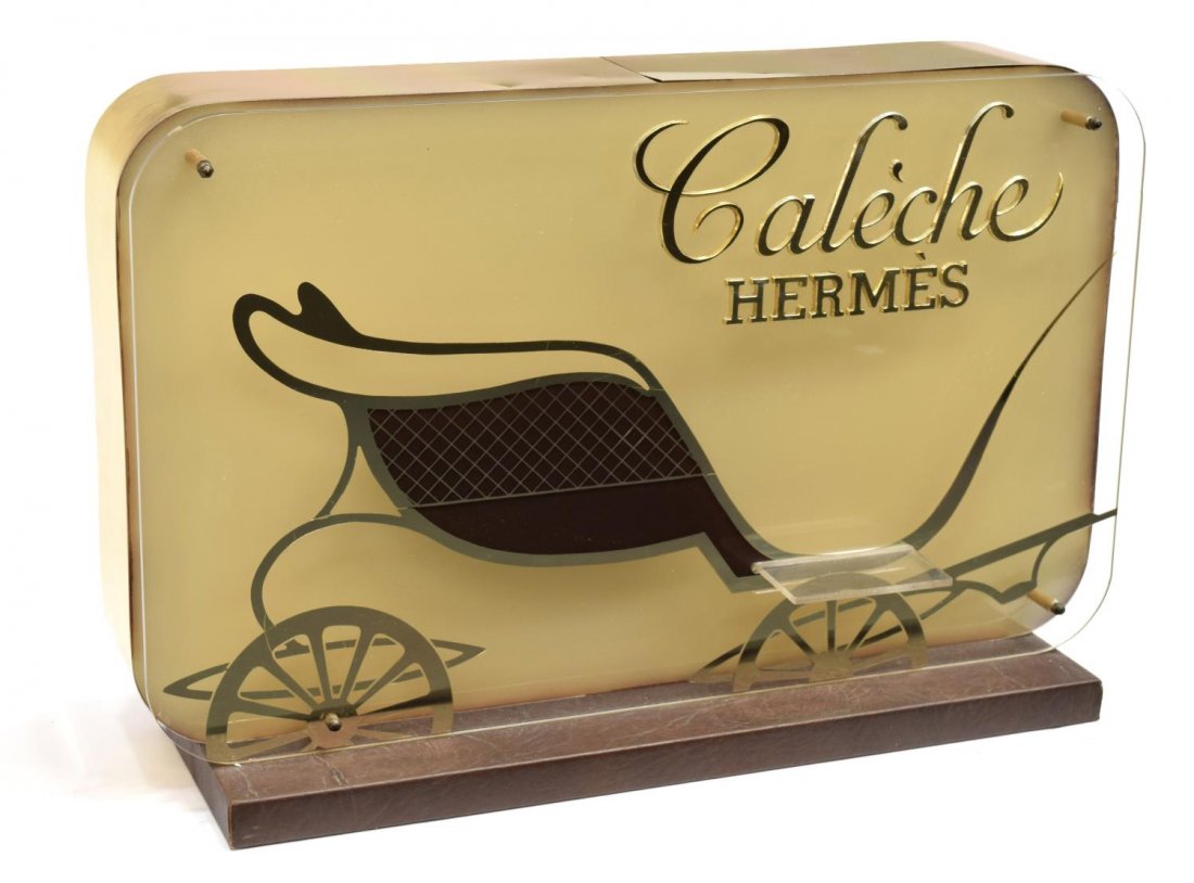 CALECHE BY HERMES ORIGINAL ADVERSTISMENT STAND: Caleche by Hermes original brass advertisement stand, 1961, plexiglass facade having gilt sled design and small clear plexiglass shelf for bottle, on vinyl wrapped base, lighted in need of wiring, 12"