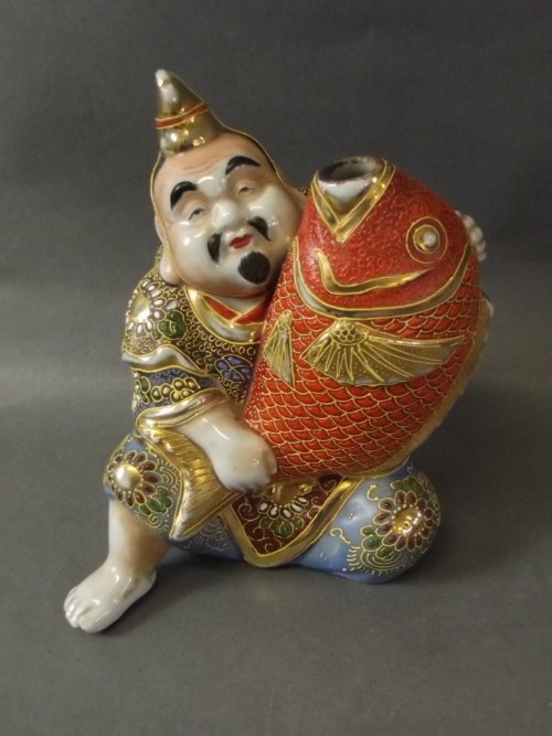 A Satsuma figure of a seated man holding a large carp,: A Satsuma figure of a seated man holding a large carp, well coloured and outlined in gilt, 6'' high