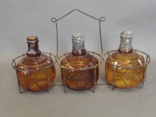 A set of 3 Hayward's Hand Fire grenades made of thick: A set of 3 Hayward's Hand Fire grenades made of thick amber glass grenades with raised markings, in original wire fire side basket, 15'' long