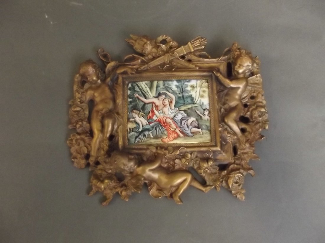 An ornate brass easel frame with winged cherub: An ornate brass easel frame with winged cherub decoration containing a cold enamel plaque depicting a classical scene, 7'' x 6''