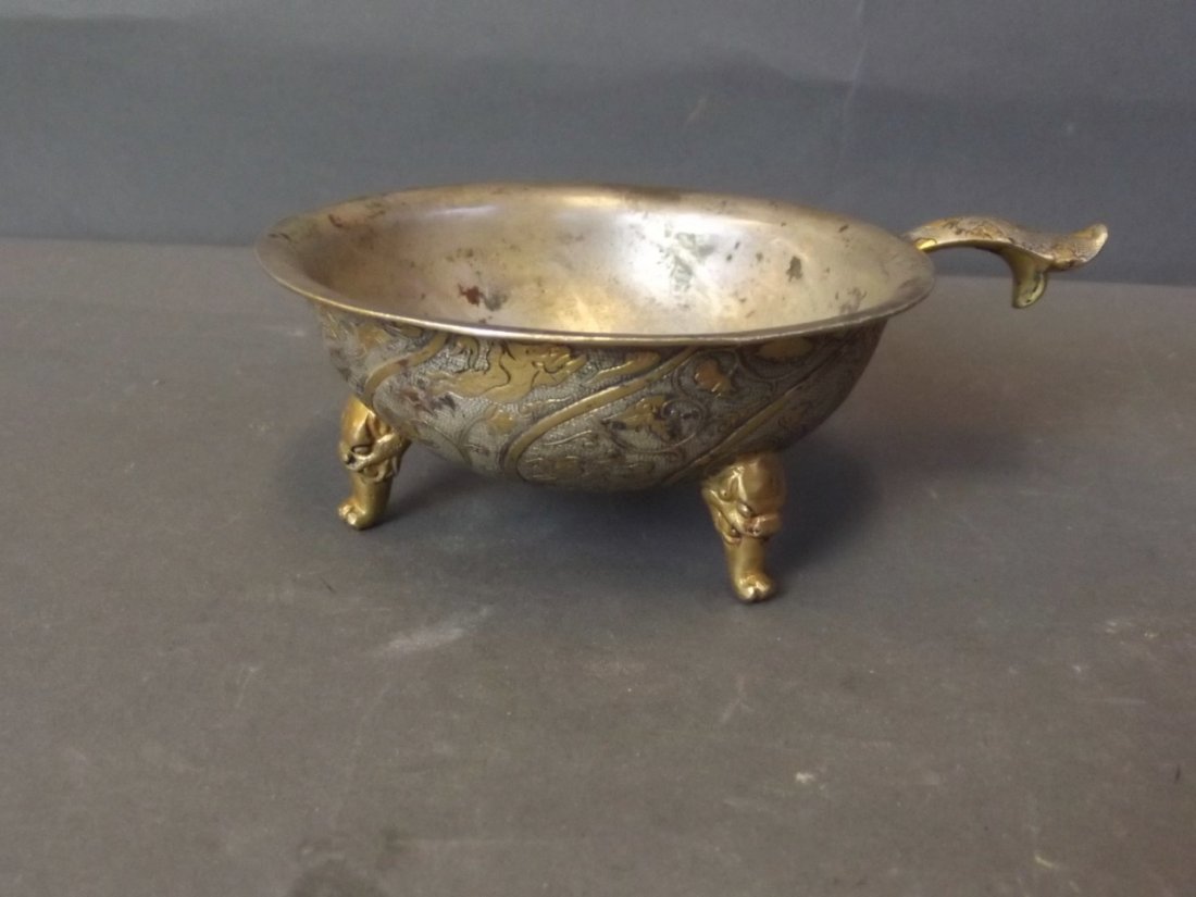 A Chinese silver plated and gilt washed bronze libation: A Chinese silver plated and gilt washed bronze libation cup, the interior with relief decoration of two fighting kylin, the exterior in stylised lotus form inset with trailing foliage, standing on tri