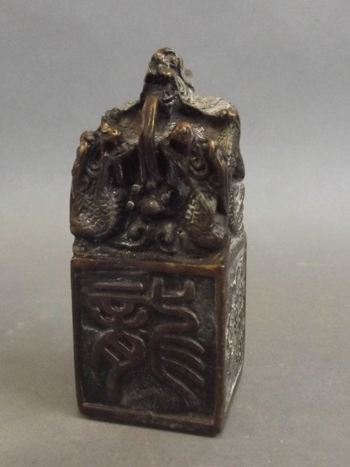 A Chinese bronze seal of square form, the top cast as: A Chinese bronze seal of square form, the top cast as three dragons, the sides decorated with symbols and calligraphy, 4½" high