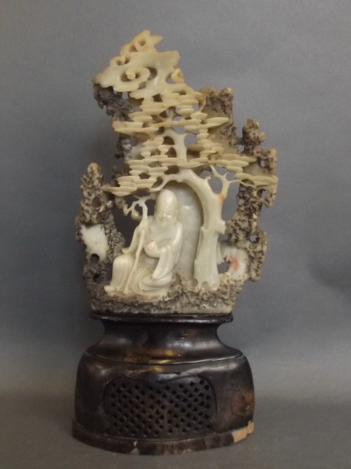 A Chinese soapstone carving of Shao Lao seated in a: A Chinese soapstone carving of Shao Lao seated in a forest, on a carved and pierced stand, 11" high