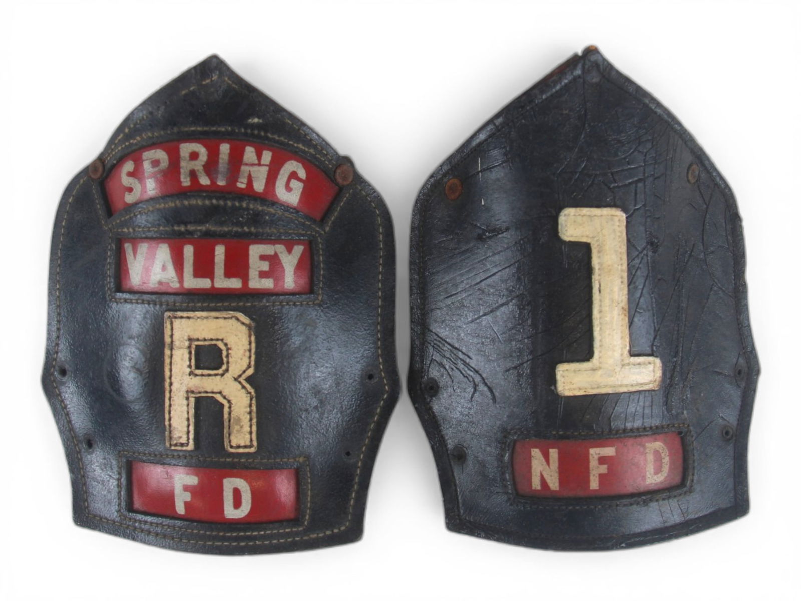(2) Vintage Leather Fire Helmet Shields: Group of two vintage leather fire helmet front shields. One shield is lettered "SPRING VALLEY" above a large letter "R," with a red "FD" panel at the base, while the second shield features a large