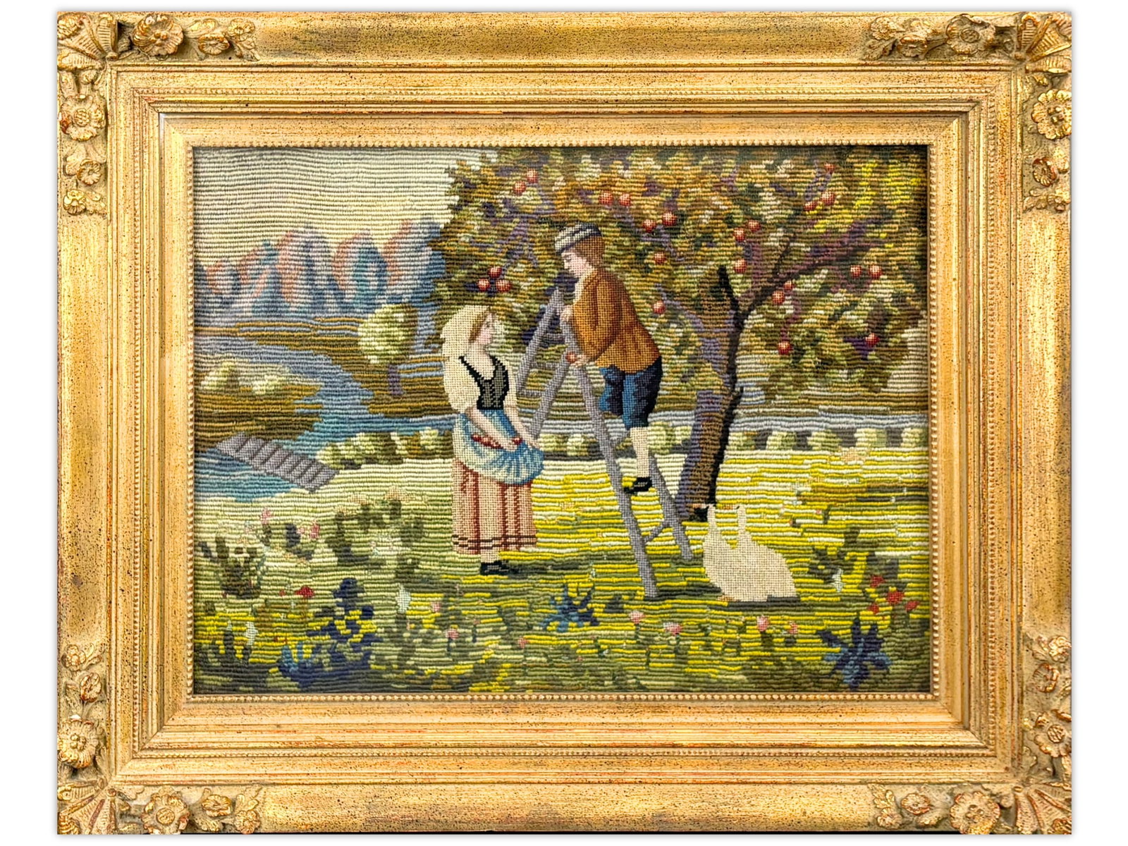 Framed Needlepoint Pastoral Scene: Hand-worked needlepoint textile depicting a charming pastoral scene with a young couple gathering apples beneath a fruit tree. The composition features a winding stream, distant mountains, colorful fl