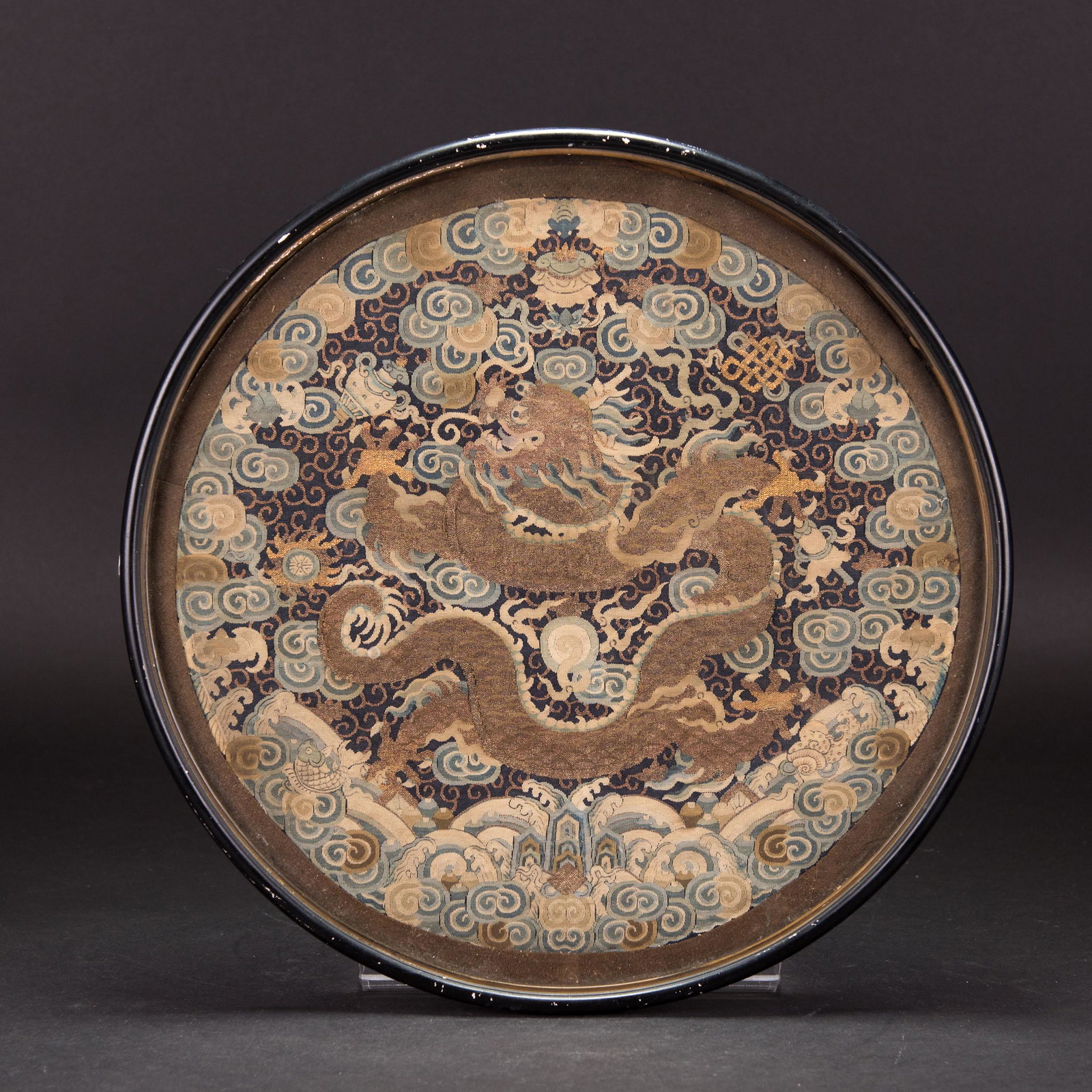 A CHINESE FRAMED DRAGON RANKING BADGE: Round, embroidered ranking badge of a dragon. Dragon is 5-clawed. It is surrounded by clouds and the 8 Buddhist Emblems. At the bottom is a wave and rock motif. It is framed in a round frame with glas