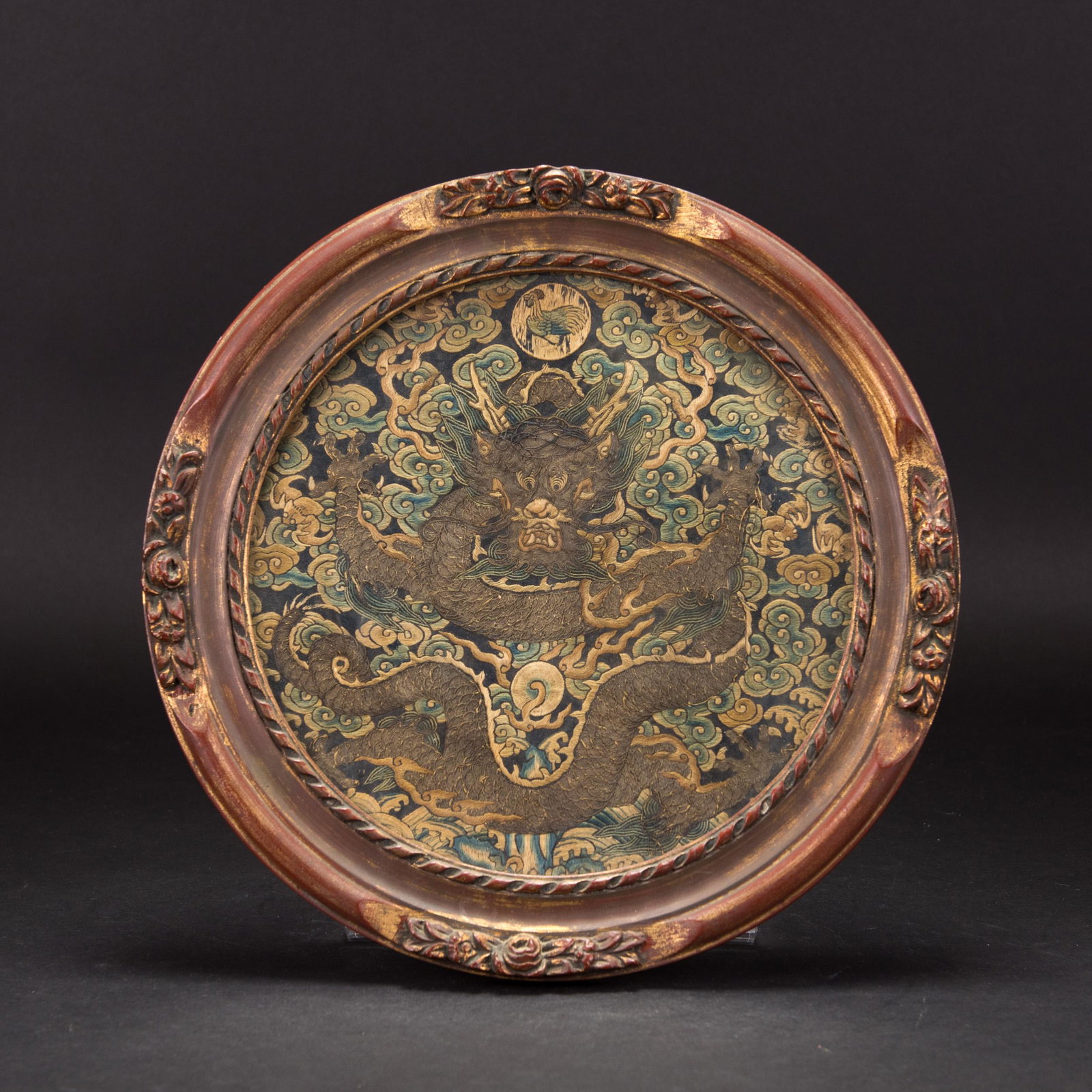 CHINESE FRAMED DRAGON RANKING BADGE: Round dragon ranking badge from the 18th century in a wooden frame. An embroidered ranking badge of a 5-clawed dragon chasing a flaming pearl. The badge is blue ground with a cloud pattern. Above the 