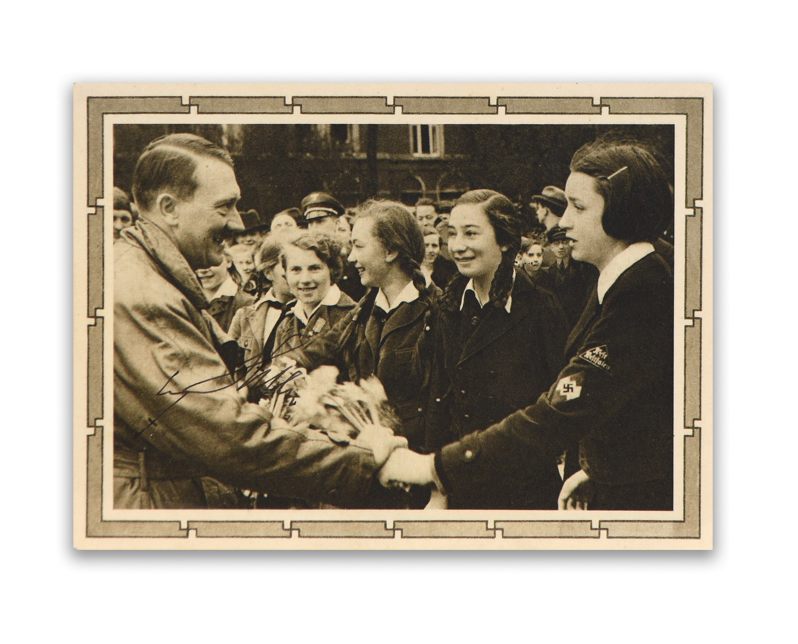 PHOTOGRAPHIC POSTCARD BEARING SIGNATURE READING 'ADOLF HITLER': A vintage photographic postcard depicting Adolf Hitler greeting a group of young women during a public appearance, with a crowd gathered in the background observing the interaction; the image captures