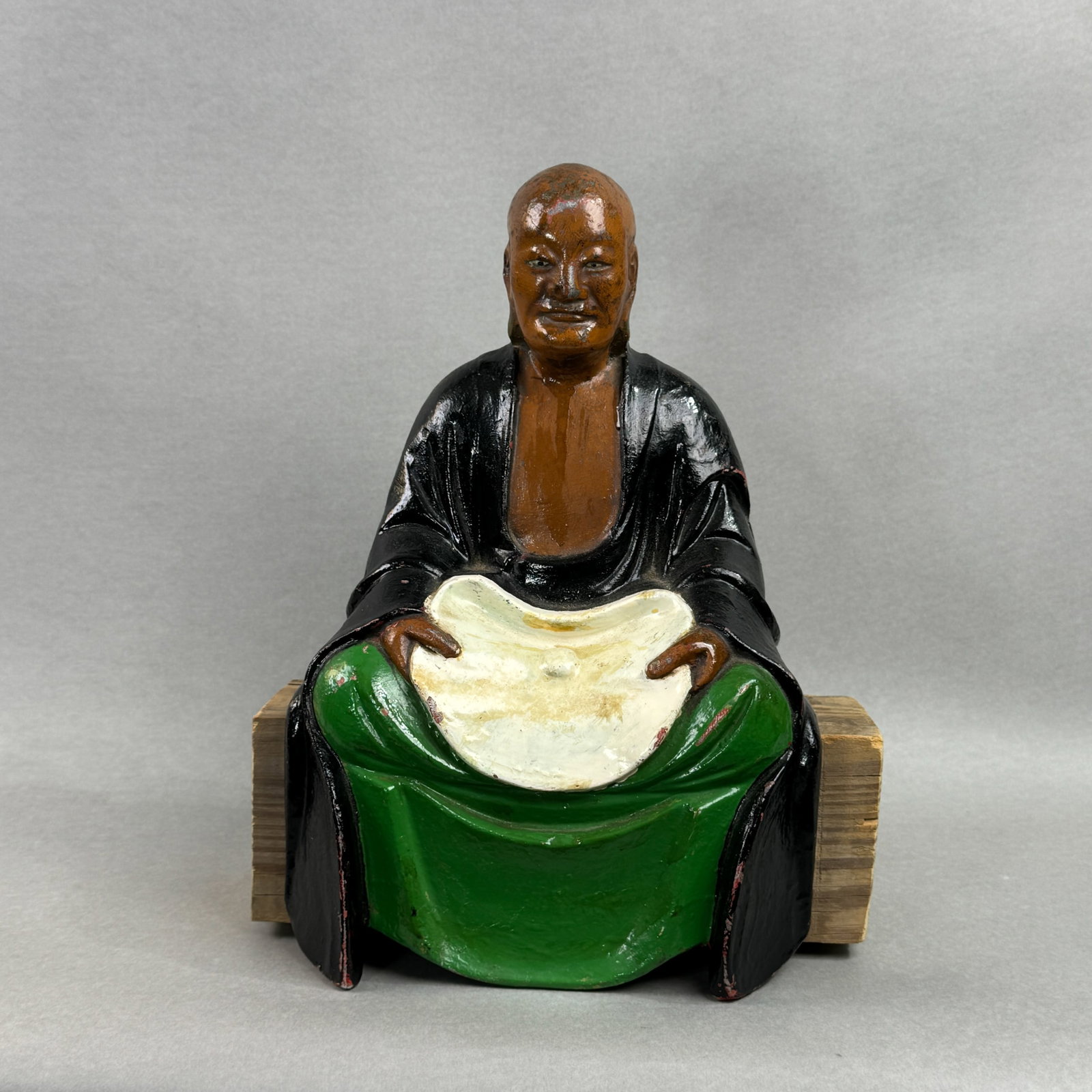 A JAPANESE IRON MONK WITH POLYCHROME PIGMENT: A Japanese iron monk with polychrome pigment, 19th/20th century. Heavy for its size, seated on a block of wood. Losses of paint. Dimensions: Height: 12.5 in. (31.8 cm); Width: 8.5 in. (21.6 cm).