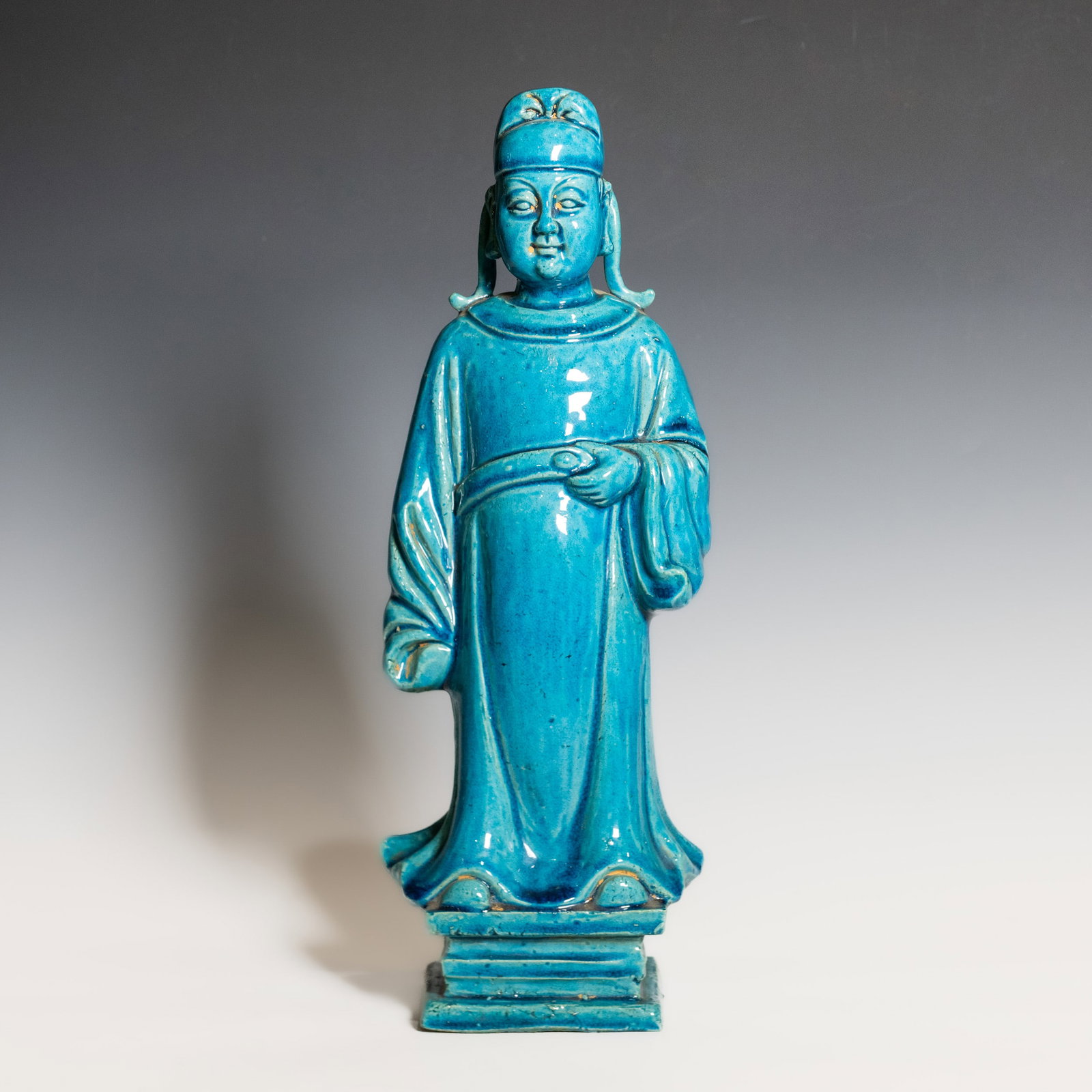 A CHINESE TURQUOISE-GLAZED CERAMIC FIGURE OF AN OFFICIAL: A Chinese turquoise-glazed ceramic figure depicting a standing official, modeled wearing a tall official's cap and flowing long robes, with both hands holding a hu tablet before the chest. The figure 