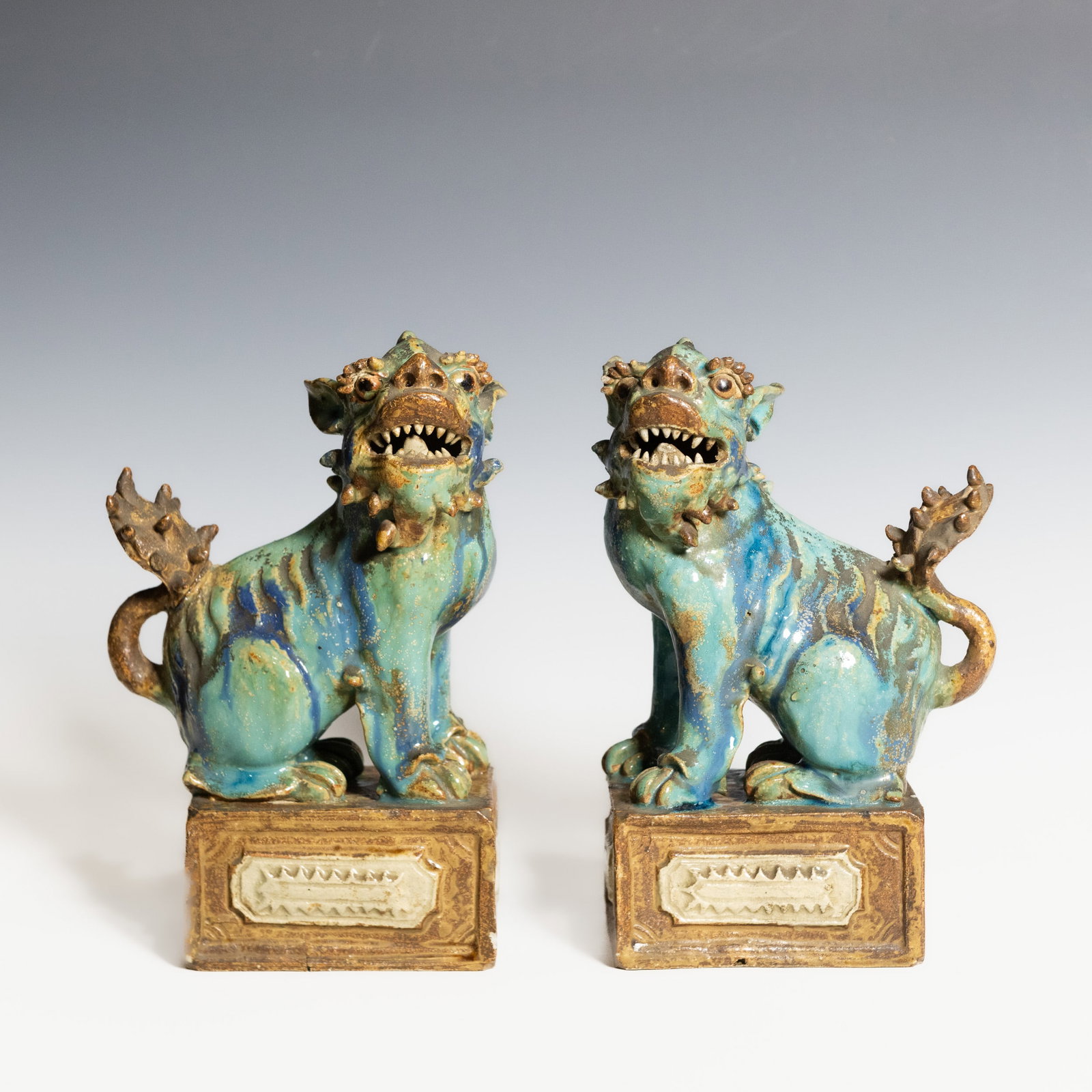 (LOT OF 2) A PAIR OF CHINESE TURQUOISE-GLAZED SHIWAN BUDDHIST LION FIGURES: (lot of 2) A pair of turquoise-glazed Shiwan Buddhist lion (foo dog) figures, each modeled seated on a rectangular plinth with an open mouth exposing sharp teeth, bulging eyes, a raised curled tail, a