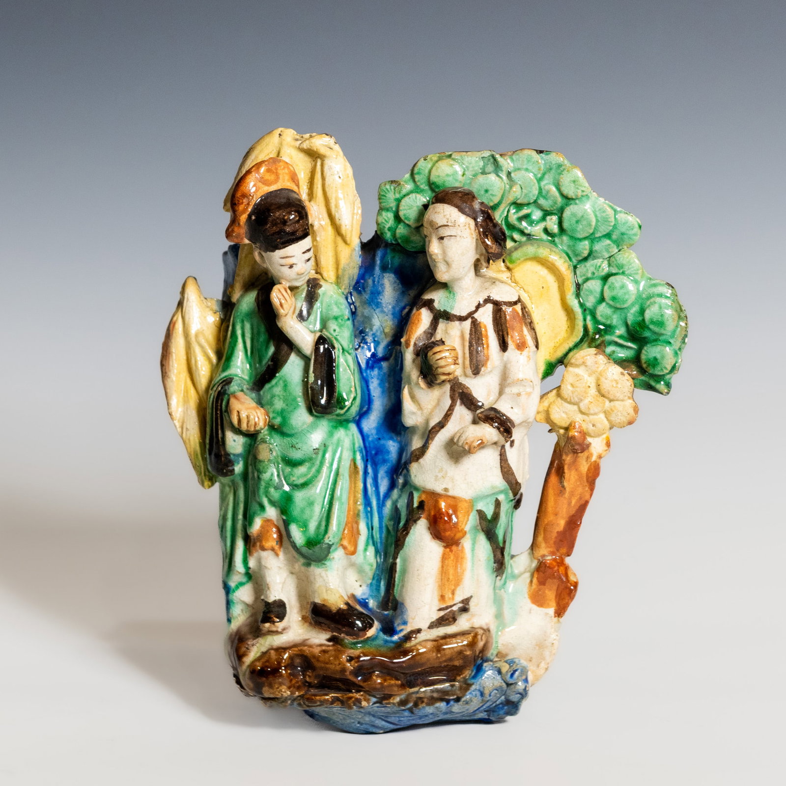 A SHIWAN POLYCHROME-GLAZED POTTERY FIGURE GROUP OF TWO SCHOLARS: A Shiwan polychrome-glazed pottery figure group of two scholars, modeled in the round, depicting two scholars standing in conversation beneath a gnarled tree. One figure is dressed in a flowing green 