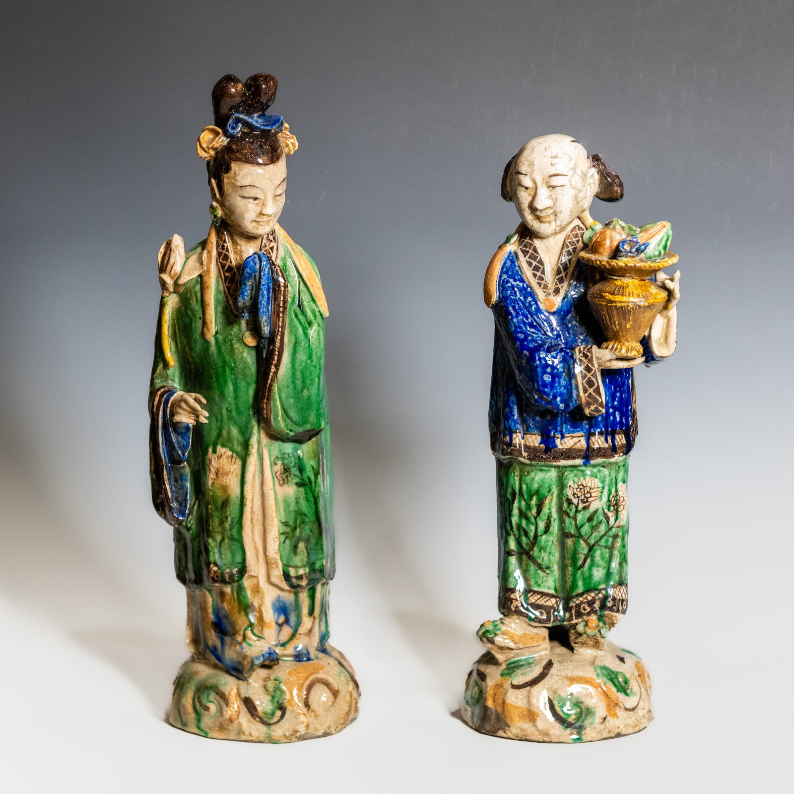 (LOT OF 2) A PAIR OF CHINESE POLYCHROME PORCELAIN FIGURES: (lot of 2) A pair of Chinese polychrome crackle-glazed porcelain figures depicting elegantly dressed female attendants. One figure holds a fruit-filled vessel while the other stands with a long-handle