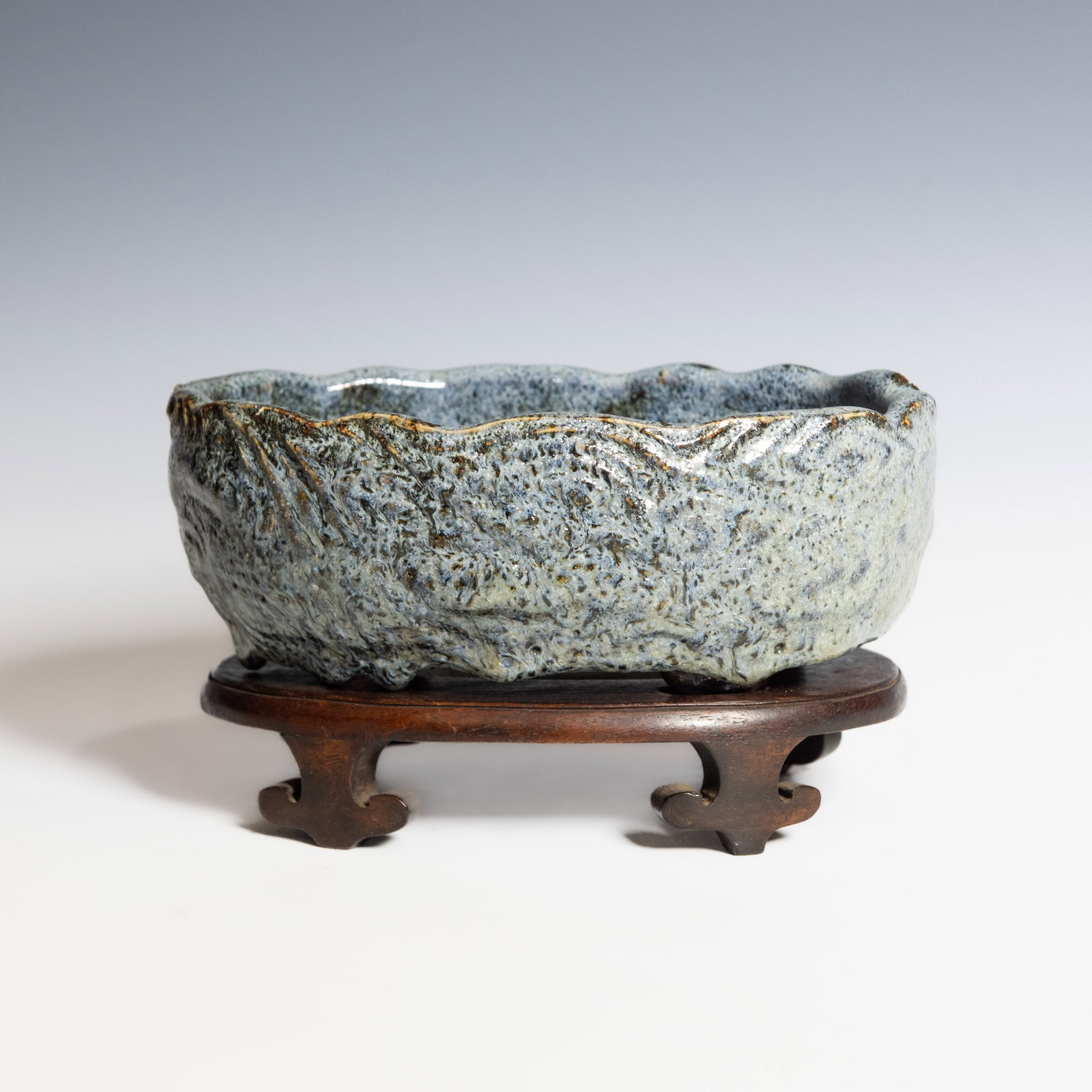 A CHINESE SHIWAN GLAZED BASIN WITH BASE, QING DYNASTY: A Chinese Shiwan glazed stoneware basin of irregular foliate form, Qing dynasty. The rim is shaped with undulating lobes, and the interior is covered in a speckled grey glaze with darker mottling. Ove