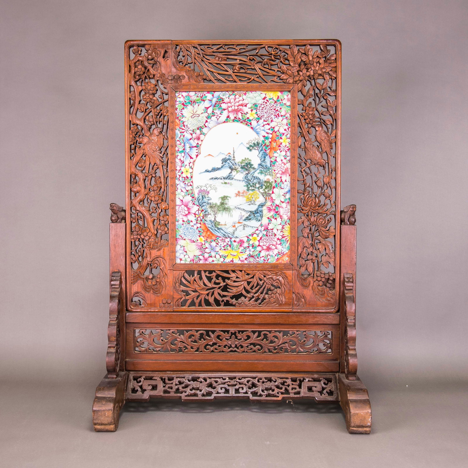 FAMILLE-ROSE MILLEFLEURS 'LANDSCAPE' TABLE SCREEN WITH ORIGINAL WOODEN STAND: A famille-rose millefleurs 'landscape' table screen with a wooden stand, 19th/20th century. The screen is painted in oval panels with landscape scenes, all against a colorful array of flowers