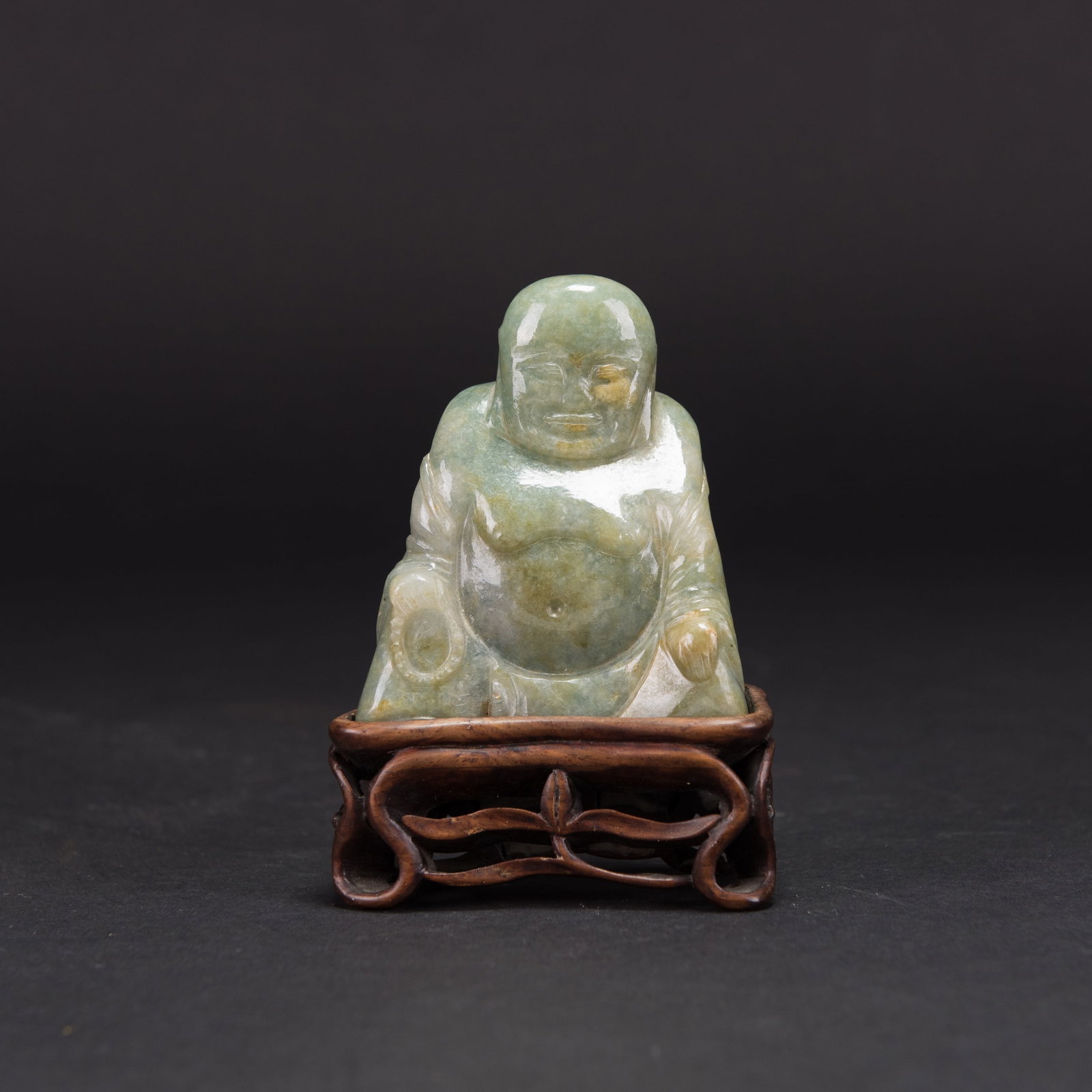 CARVED JADEITE BUDDHA: Carved jadeite seated Buddha with rust-colored suffusions, on a wood base. Overall (with base): 4.25"h x 1.5"w x 2.75"l (10.8 x 3.8 x 7 cm)