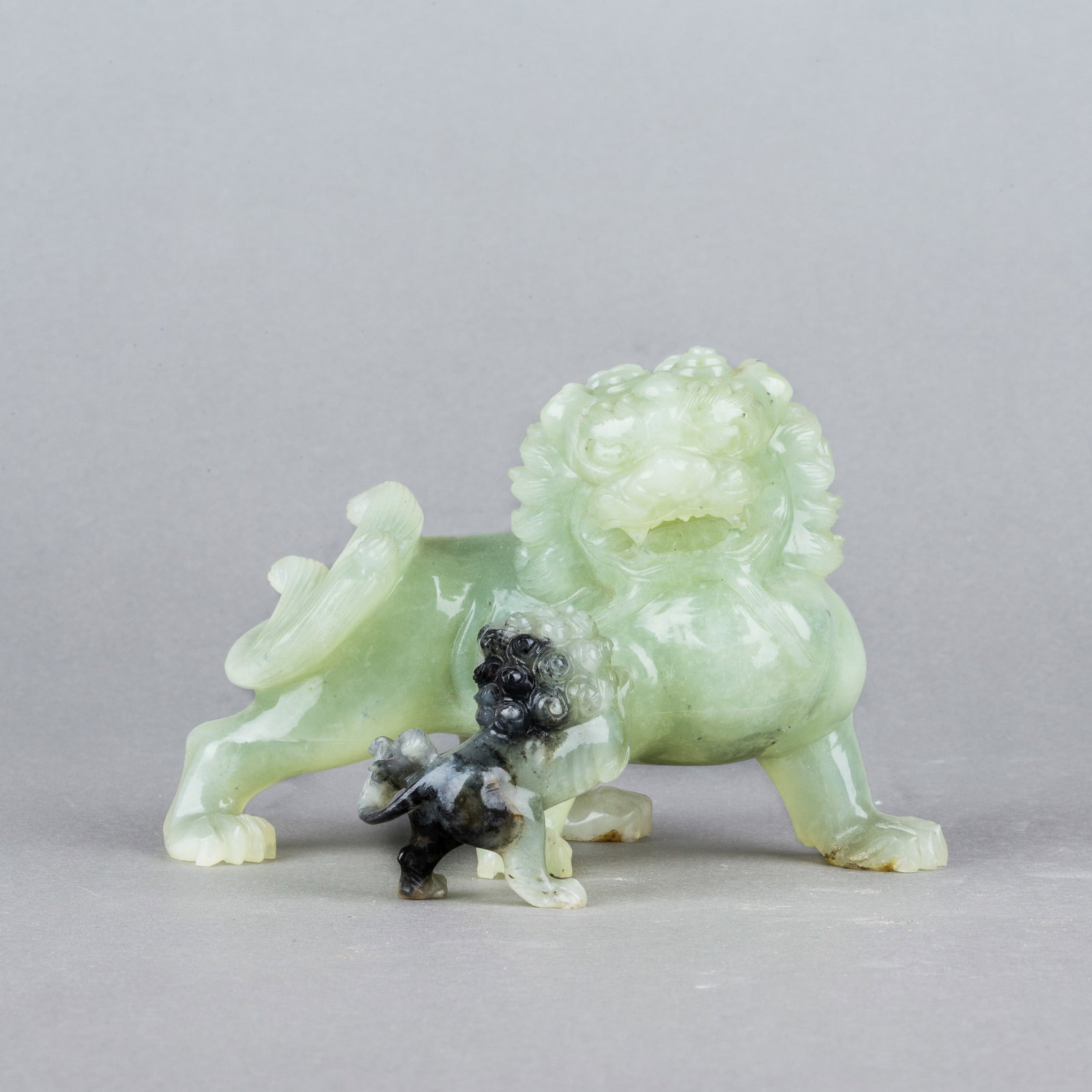 (LOT OF 2) GROUP OF TWO JADE LIONS: (lot of 2) A group of two jade lion carvings, one large and one small. The stone is of a light green tone, with dark russet coloration on the smaller lion. H9 x 14 cm