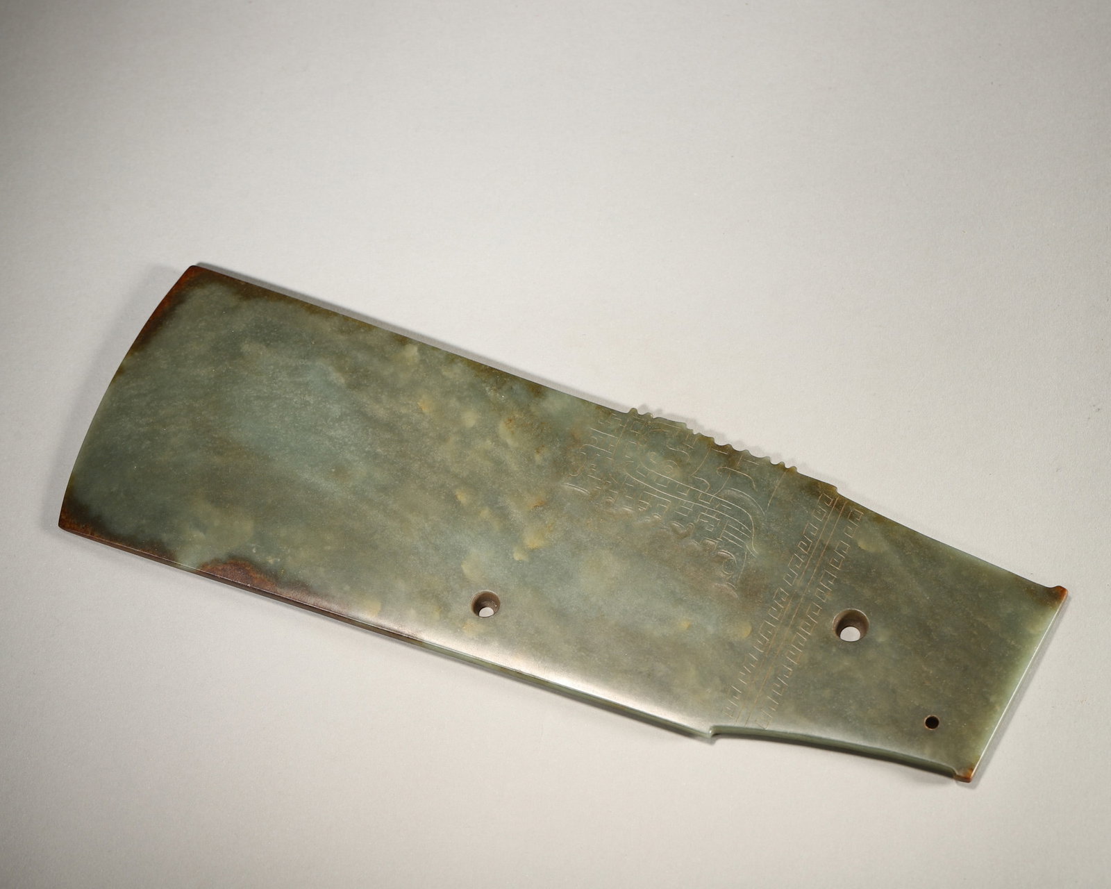 A GREEN JADE BLADE-FORM RITUAL OBJECT: A flat green jade blade-form ritual object carved with beast-mask and geometric motifs. L. 30 cm; W. 9.5 cm; Wt. 470 g.