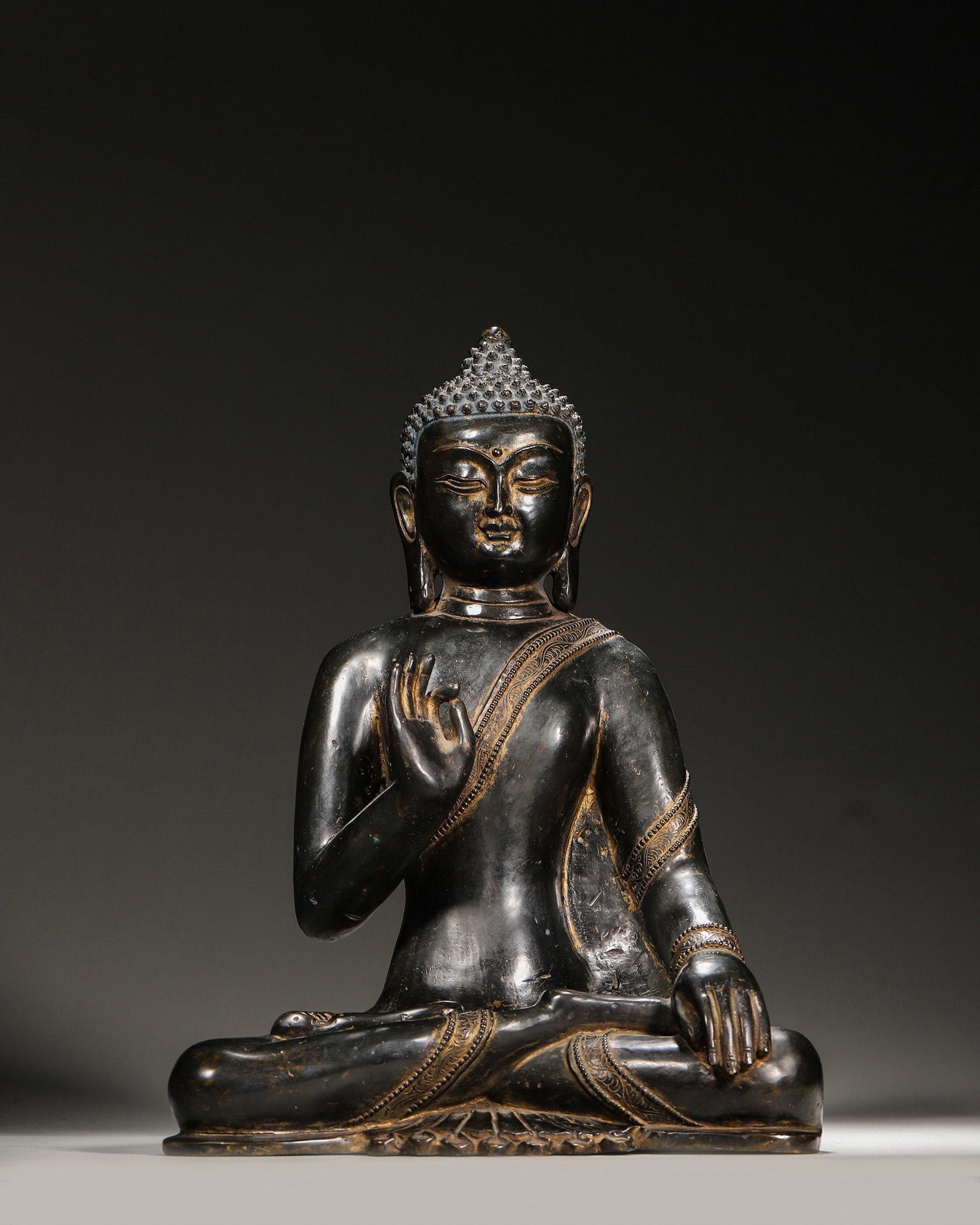 A BRONZE FIGURE OF DIPANKARA BUDDHA: A bronze figure of Dipankara Buddha seated on a lotus base, the right hand raised in abhaya mudra and the left hand resting on the knee, with patterned robe borders and a vajra motif to the base. L.