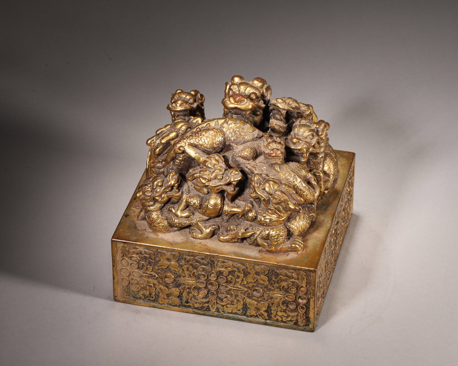 A GILT-BRONZE NINE-DRAGON SEAL: A gilt-bronze seal surmounted by a nine-dragon finial with cloud motifs, the body decorated with floral patterns. The seal face bears seal-script characters. L. 15 cm; W. 15 cm; Ht. 14 cm; Wt. 15.85 k