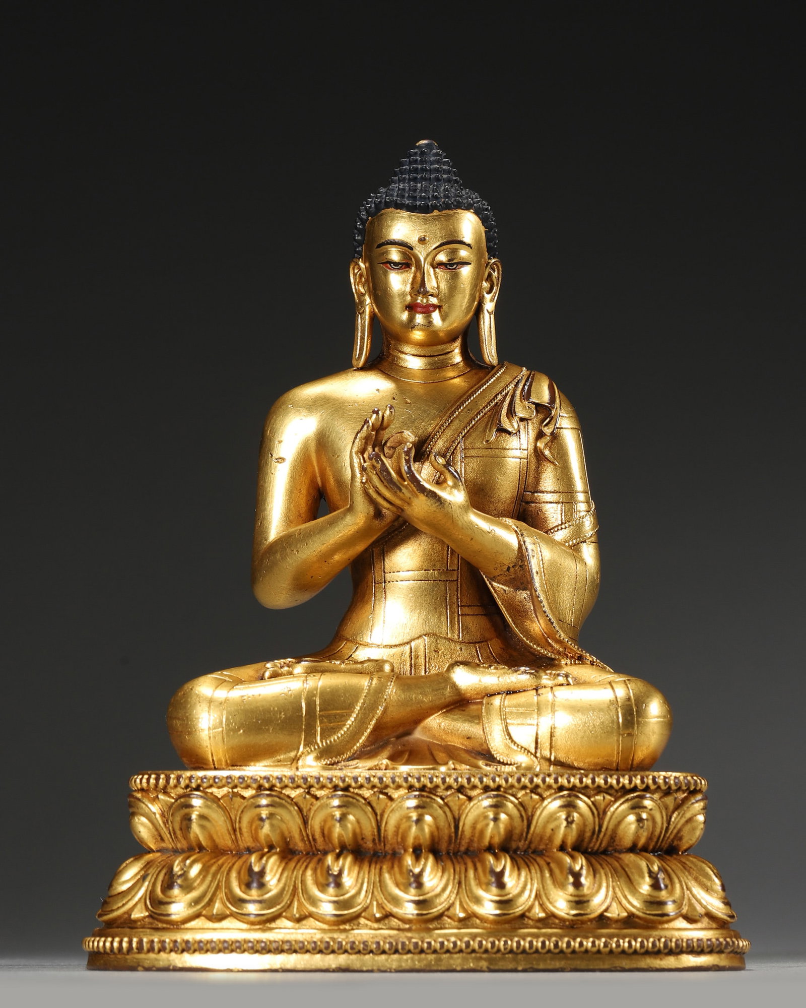 A GILT-BRONZE FIGURE OF SHAKYAMUNI BUDDHA: A gilt-bronze figure of Shakyamuni Buddha seated on a lotus base, with dark blue hair, patterned robes and hands held in dharmachakra mudra. W. 12 cm; Ht. 16 cm; Wt. 859 g. Note: By repute, in Tibetan