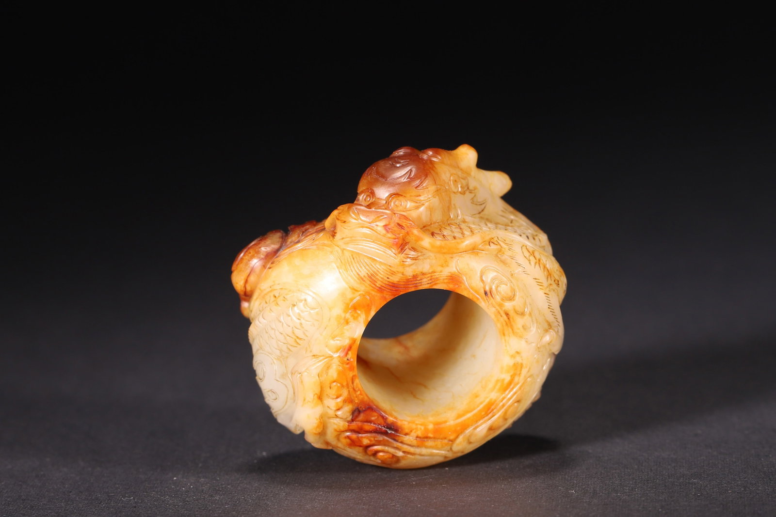 A JADE THUMB RING WITH DRAGON CARVING: A jade thumb ring carved in high relief with a dragon and cloud motifs, with areas of russet skin. L. 5.3 cm; W. 5 cm; Ht. 3.7 cm; Wt. 101 g.