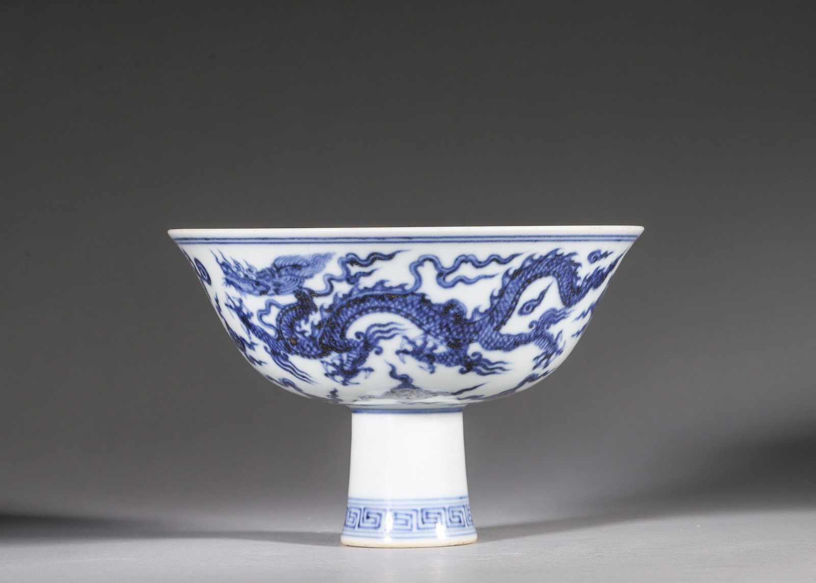 A BLUE-AND-WHITE 'DRAGON' STEM BOWL: A blue-and-white porcelain stem bowl decorated with two dragons pursuing flaming pearls amid clouds, with a key-fret band to the stem. The interior bears a six-character Xuande mark within double circ