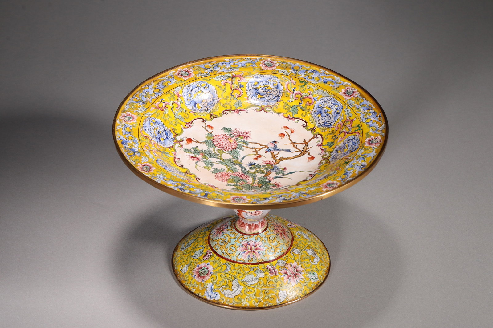 A COPPER-BODY PAINTED-ENAMEL YELLOW-GROUND STEM DISH WITH BIRDS AND FLOWERS: A copper-body painted-enamel stem dish with a gilt rim, decorated with birds and flowers within a central reserve and floral scrolls on a yellow ground. The stem is decorated with bats and clouds,