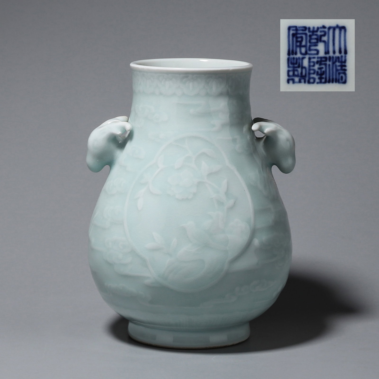 A LIGHT-CELADON-GLAZED VASE WITH DEER-HEAD HANDLES AND FLORAL PANELS: A porcelain vase covered in a light celadon glaze, with deer-head handles and carved floral and bird panels. The base bears a mark described in the source as Qianlong. W. 13.5 cm; Ht. 19 cm; Wt. 1 kg.
