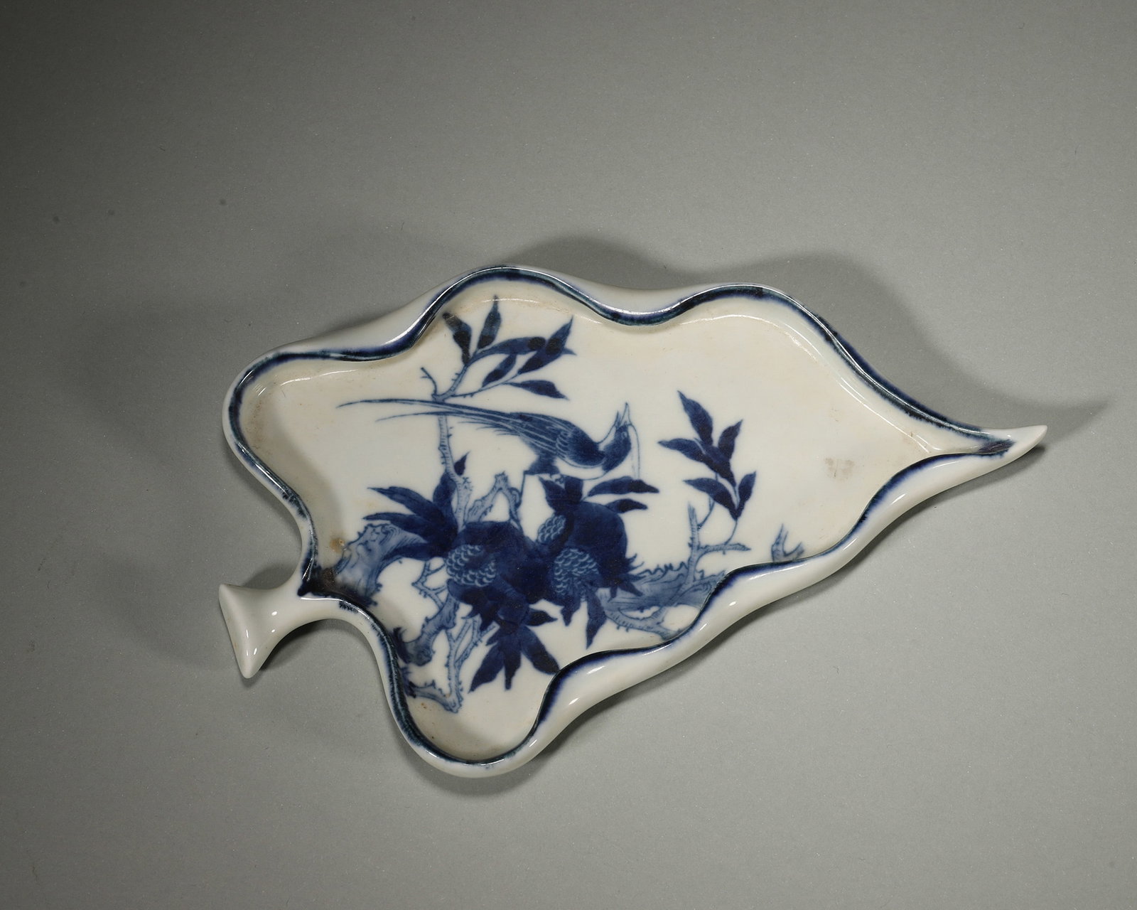 A BLUE-AND-WHITE LEAF-FORM INK PALETTE WITH BIRD AND FRUIT: A blue-and-white porcelain leaf-form ink palette decorated with a bird and fruiting branches. L. 15.8 cm; W. 7.5 cm; Wt. 111.1 g.