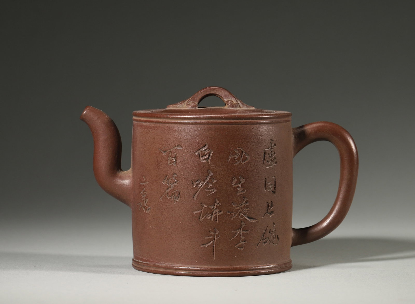 A ZISHA CYLINDRICAL TEAPOT WITH POEM INSCRIPTION: A zisha teapot with a straight cylindrical body, bridge-form knob, spout and handle, with a carved calligraphic poem to the body. L. 13 cm; W. 7.5 cm; Ht. 8.5 cm.