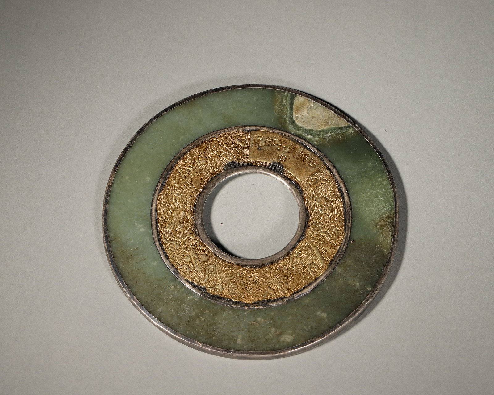 A JASPER BI WITH SILVER MOUNT AND GILT EIGHT TREASURES: A jasper bi with a silver rim, one side bearing a gilt poem inscription, the inner ring decorated in gilt with the Eight Treasures and cloud motifs. W. 14.2 cm; Wt. 299 g. Note: By repute, remounted f