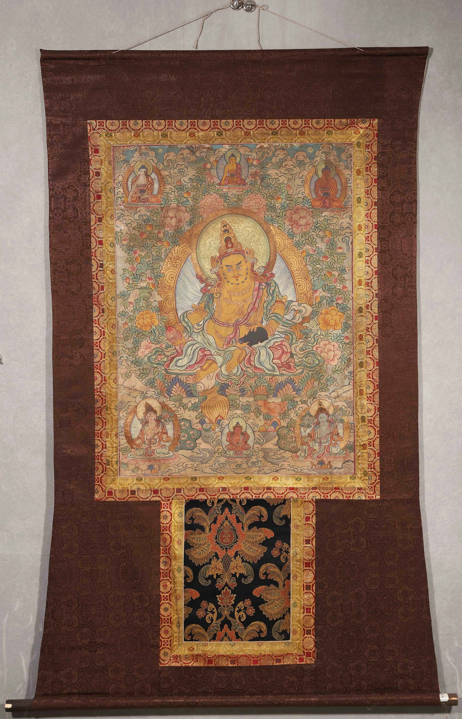 A THANGKA OF YELLOW JAMBHALA: A mineral-pigment thangka depicting Yellow Jambhala seated in royal ease, holding a mongoose and a gem, with Buddhist figures arranged above and around the central deity, with gold details. The